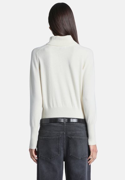 Femme aux longs cheveux foncés portant un pull à col roulé blanc et un jean gris foncé taille haute avec une ceinture noire, vue de dos.