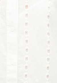 Λευκό ύφασμα με κάθετα κεντημένα σχέδια αλυσίδας και κουμπιά κατά μήκος της αριστερής πλευράς.