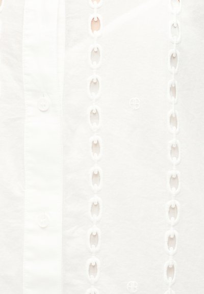 Hvidt stof med lodrette broderede kædemønstre og knapper langs venstre side.