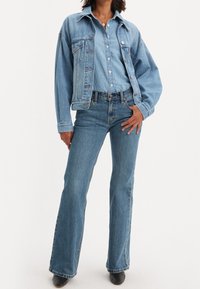 Veste en denim bleu clair, chemise assortie et jeans évasés. La veste comporte des poches avant et une fermeture à boutons. Denim texturé avec légère décoloration.