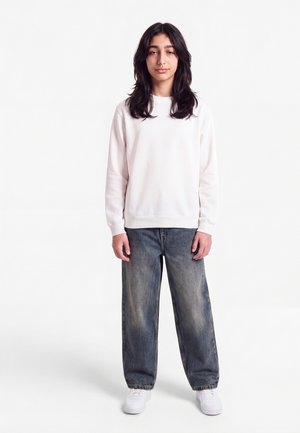 Jeune personne aux longs cheveux foncés portant un sweat-shirt blanc, un jean délavé à jambes larges et des baskets blanches, debout sur un fond blanc.