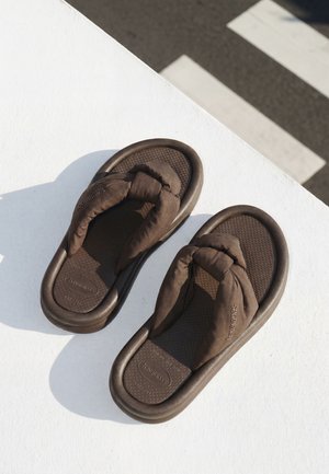 Sandales tongs marron rembourrées avec des brides matelassées posées sur une surface blanche à côté d’un passage piéton ombragé.