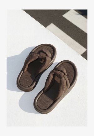 Smeđe flip-flop sandale s jastučićima i podstavljenim trakama postavljene na bijelu površinu pokraj zasjenjenog pješačkog prijelaza.