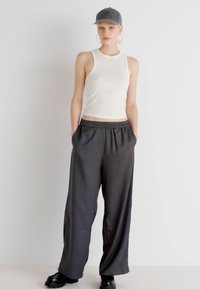 Witte geribde tanktop en losse donkergrijze broek met een elastische tailleband. Zwarte schoenen en een grijze pet. Minimalistisch ontwerp en ontspannen pasvorm.