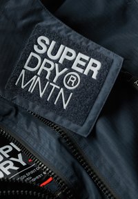 Interior de una chaqueta de navy con un parche texturizado negro que lleva el texto en relieve blanco "SUPER DRY® MNTN." Incluye una cremallera visible y una tela con patrón en cuadrícula.