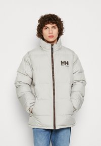 Pufová bunda vyrobená z lehkého šedého materiálu s vysokým límcem. Hnědý zip a značka na hrudi. Obsahuje prošívaný design.