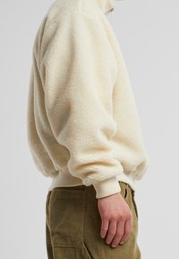 Cremfarbener Fleece-Pullover mit strukturierter Oberfläche, hohem Kragen und gerippten Bündchen, kombiniert mit khakifarbenen Cordhosen.