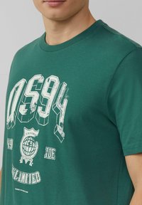 Groene T-shirt van zachte katoen, voorzien van een grafische print in het wit, inclusief "QS94" en een wereldbol-embleem. Ronde halslijn en korte mouwen.