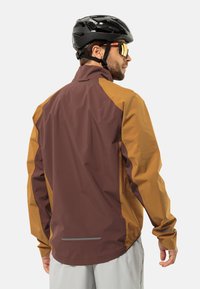 Veste de cyclisme imperméable avec un corps brun et des accents moutarde, un col montant, des manches longues et une bande réfléchissante à l'arrière.