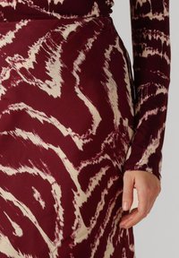 Bordeauxrotes Kleid mit einem strukturierten, abstrakten Creme-Muster. Lange Ärmel und ein figurbetonter Schnitt, der die Taille betont. Dezenter Glanz des Stoffes.