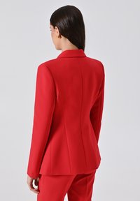 Donna dai lunghi capelli scuri che indossa orecchini d'oro e un blazer rosso su misura abbinato ai pantaloni, vista da dietro su uno sfondo semplice.