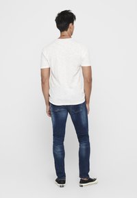 Weißes T-Shirt mit kurzen Ärmeln, kombiniert mit dunkelblauen Denim-Jeans. Die Jeans sind schmal geschnitten und weisen subtile Verfärbungen auf, und das Modell trägt schwarze Sneakers.