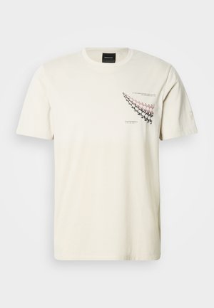 T-shirt en coton couleur crème à manches courtes, avec un motif graphique de flèches en rose et noir sur le côté gauche de la poitrine.