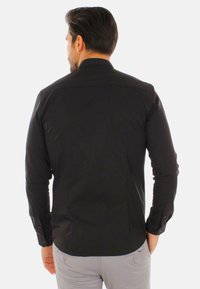 Camicia nera a maniche lunghe con colletto standard, superficie liscia, cuciture laterali e polsini con bottoni, vista dal retro.