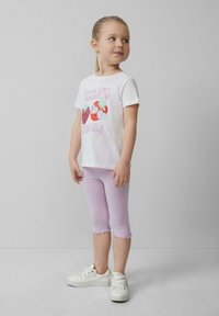 Witte t-shirt met kleurrijke fruitafbeelding en tekst, gecombineerd met pastel paarse capri leggings en witte sneakers. Glad materiaal en afgeronde zomen.