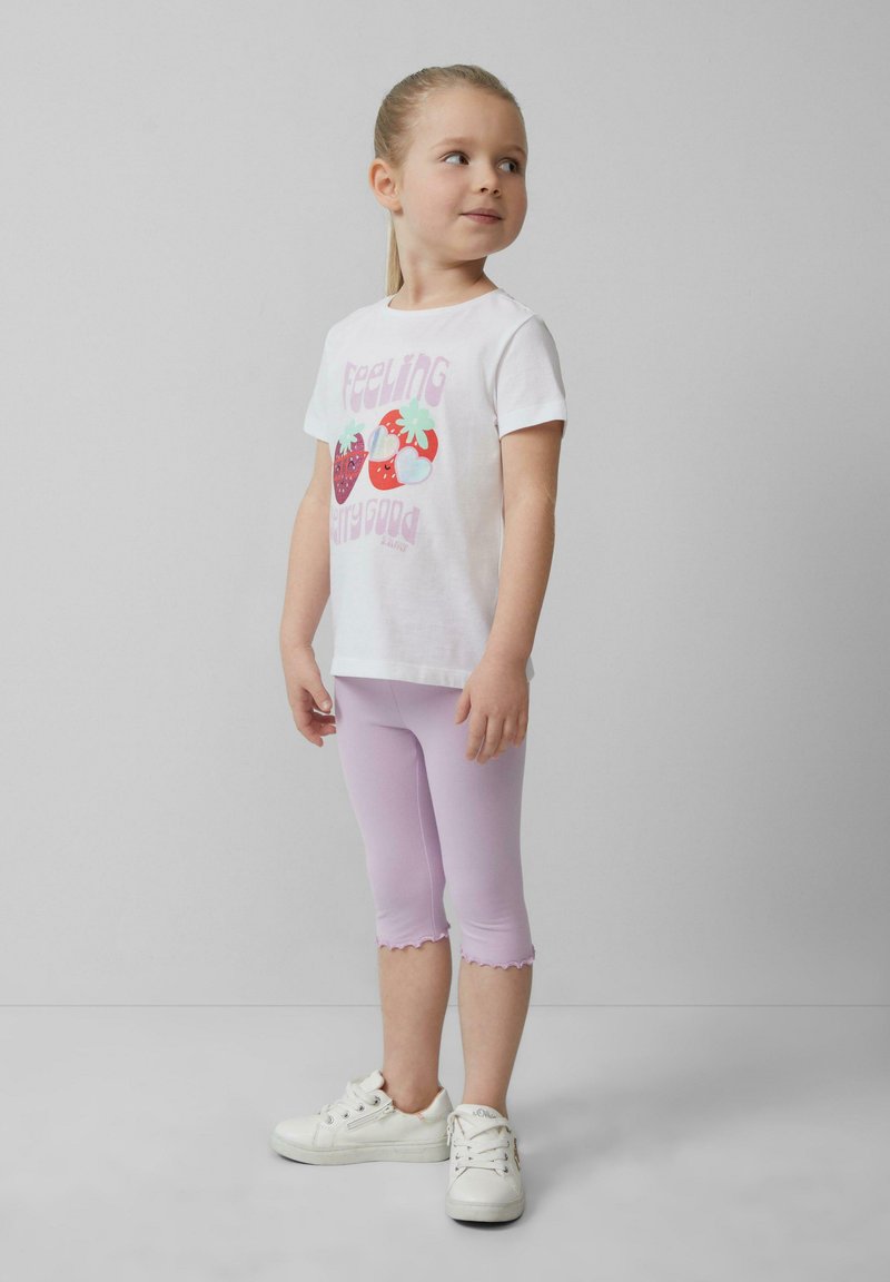 Witte t-shirt met kleurrijke fruitafbeelding en tekst, gecombineerd met pastel paarse capri leggings en witte sneakers. Glad materiaal en afgeronde zomen.