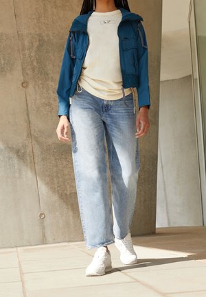 Personne portant une veste zippée bleue, un sweat crème, un jean bleu clair et des baskets blanches, debout à l'intérieur près d'un mur en béton.