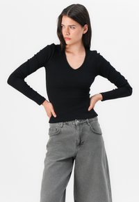 Junge Frau in einem schwarzen Langarmoberteil und grauen, weiten Jeans, die mit Händen in der Hüfte nachdenklich zur Seite blickt, vor einem weißen Hintergrund.