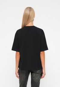 Femeie cu părul lung blond, îmbrăcată cu un tricou negru lejer și pantaloni negri texturați, stând cu spatele la un fundal alb.