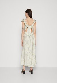 Robe maxi florale avec des bretelles volantées, dos ouvert et un détail à nœud. Tissu crème léger avec des motifs verts et jaunes. Portée avec des chaussures à talons carrés noires.