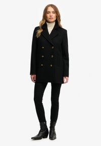 Zwarte dubbele-breasted jas met gouden knopen, piekrevers, twee voorzakken en een gestructureerde pasvorm. Draag het over een crèmekleurige coltrui.