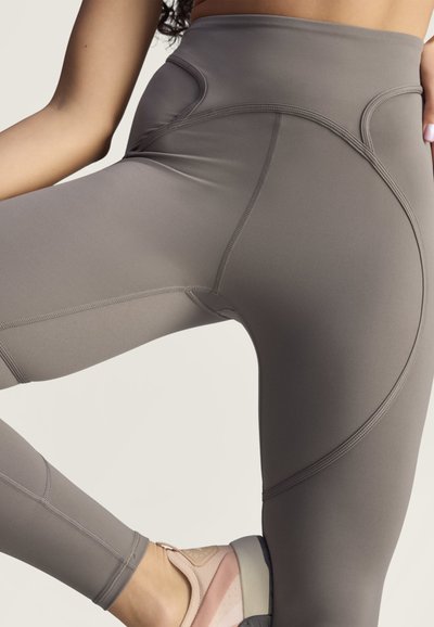 Person wearing high-waisted gray leggings and pink sneakers, lifting one leg with hand resting on the hip, against a plain background.