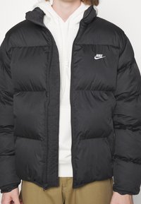 Persona vistiendo una sudadera blanca con capucha, una chaqueta de plumas Nike negra con cremallera y pantalones marrón claro, mostrada de pecho a caderas.