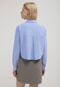 Donna con capelli biondi corti che indossa una camicia a maniche lunghe di blu chiaro e una gonna a quadretti marrone, vista di spalle su uno sfondo semplice.