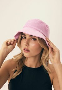 Różowa kapelusz z szerokim rondem wykonany z teksturowanej tkaniny, charakteryzujący się luźnym krojem. Modelka dostosowuje rondo w naturalnej pozie.