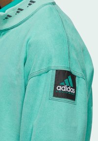 Tyrkysová mikina s žebrovaným límcem, texturovanou tkaninou a nášivkou loga Adidas v černé a tyrkysové barvě na levém rukávu.