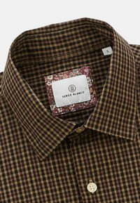 Chemise à carreaux brune et noire en tissu doux, avec un col boutonné et une étiquette à motif floral sur le col intérieur.