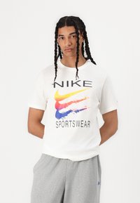 Bela bombažna majica z barvitim logotipom Nike in grafičnim oblikovanjem. V kombinaciji s sivimi trenirkami, ki prikazujejo sproščen športni slog.
