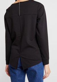 Černý top s dlouhým rukávem, vysokým výstřihem a zipem na střední zadní části. Má rozdělený lem a hladkou texturu, kombinovaný s modrými džíny.