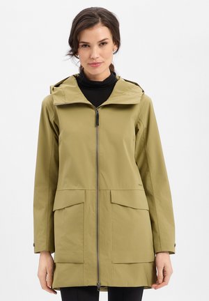 Frau trägt einen hellolivgrünen Kapuzenregenmantel mit Fronttaschen über einem schwarzen Rollkragenpullover, steht vor einem einfarbigen Hintergrund.