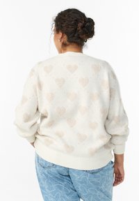 Krēmkrāsas džemperis ar reljefainu sirds rakstu gaismas bēšā krāsā. Mīksts materiāls, brīvs siluets ar ribām piedurknēs un apakšmalā. Valkāts kopā ar gaiši ziliem džinsiem.