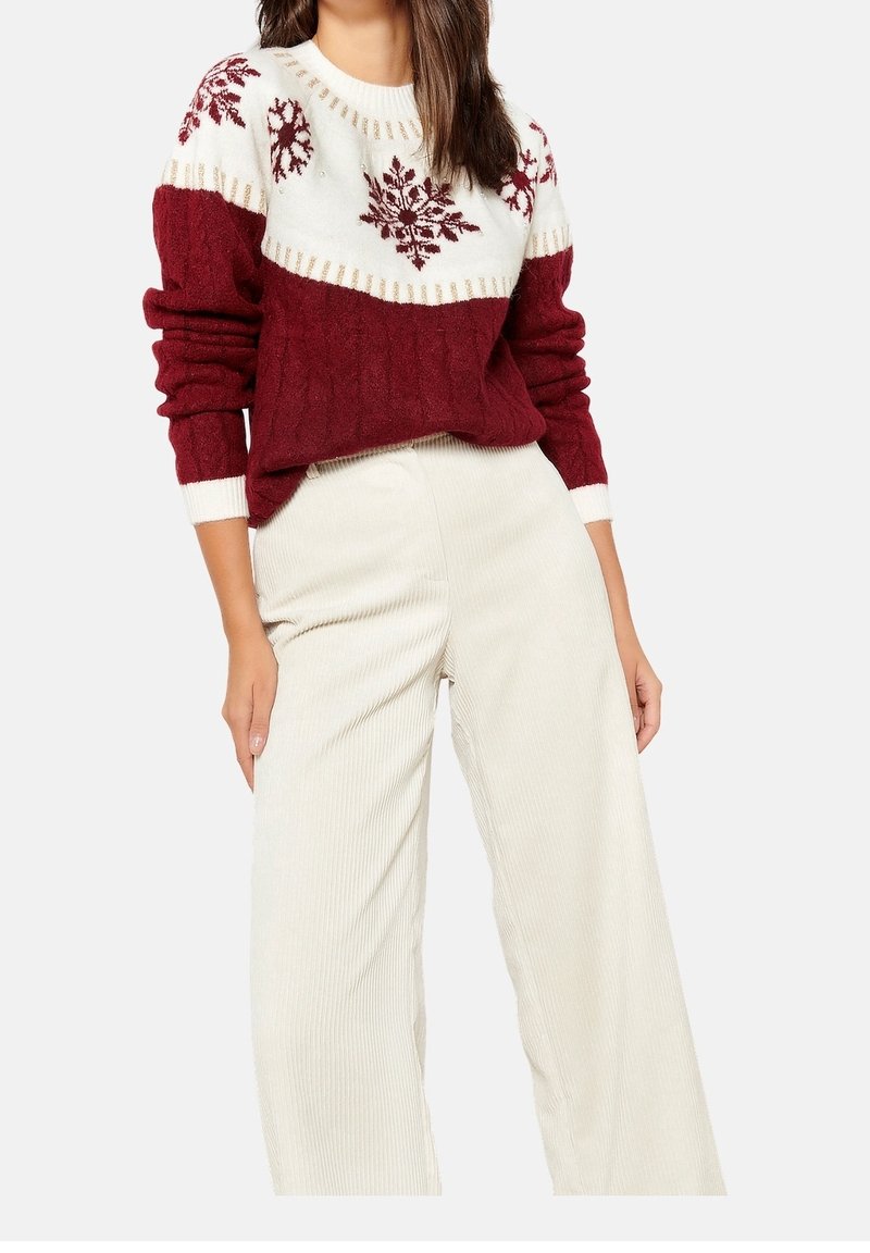 Femme portant un pull en maille bordeaux et blanc avec des motifs flocons de neige et un pantalon ample taille haute couleur crème.