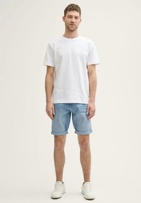 Hvid bomulds-t-shirt kombineret med lyseblå denimshorts, der er rullet op i kanten, og hvide sneakers med teksturerede snørebånd, stående på en ensfarvet baggrund.