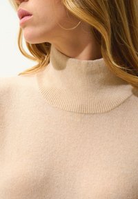 Beige Rollkragenpullover aus weichem Stoff; hat einen hohen gerippten Kragen und eine glatte, leichte Textur.