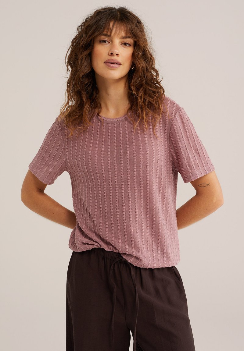 Mujer con cabello rizado que lleva una blusa corta texturizada de color malva y pantalones negros holgados, de pie con las manos detrás de la espalda.