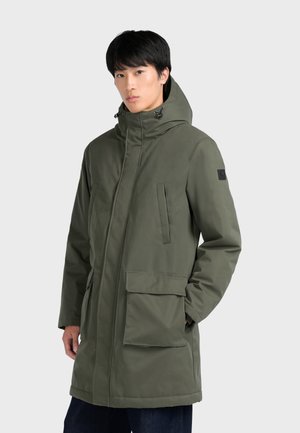 Jeune homme portant un manteau d'hiver à capuche vert olive avec de grandes poches avant, debout avec une main dans la poche sur un fond uni.