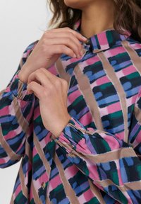 Camicia a bottoni colorata con un motivo geometrico nei toni del rosa, verde e blu. Il tessuto è morbido, con maniche a sbuffo e colletto.
