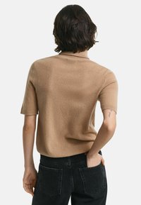 Person mit kurzen dunklen Haaren, der einen kamelfarbenen kurzärmeligen Pullover und schwarze Jeans trägt und mit einer Hand in der hinteren Tasche steht.