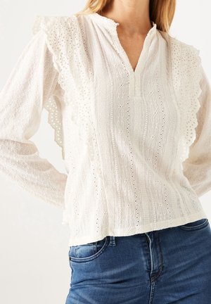 Femme portant un chemisier en dentelle blanche avec des bords festonnés et un motif texturé, associé à un jean en denim bleu.