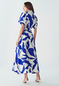 Maxi šaty s modrými listovými vzory na krémovém pozadí, krátké rukávy, s páskem v oblasti pasu a volnou siluetou. Bílé sandály na podpatku.