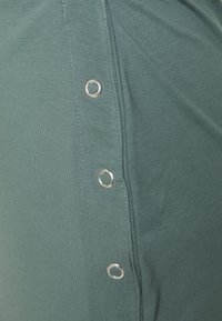 Vestido de algodão verde visto de lado, com ilhós prateados ao longo da costura, exibindo uma textura suave e um design ajustado.