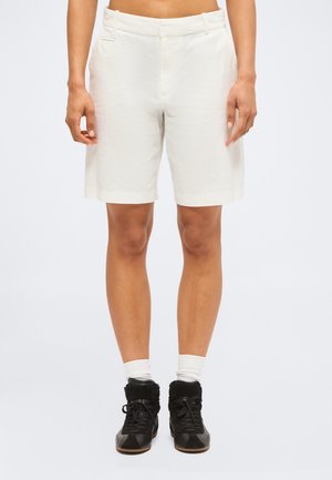 Osoba o szczupłych nogach, ubrana w białe szorty z wysokim stanem, białe skarpety i czarne sneakersy na sznurowadła, stojąca na tle jednolitego tła.