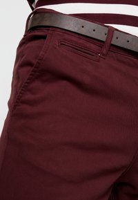 Burgunderfärgade bomullsbukser med smal passform, med en slät frontficka och ett brunt läderbälte, strukturerad tyg med lätt stretch.