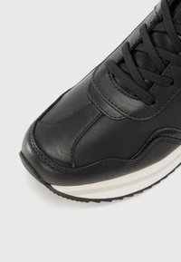 Baskets en cuir noir avec une pointe arrondie, surface texturée et lacets plats. Elles présentent une semelle intermédiaire blanche avec trois rayures noires visibles sur la semelle extérieure.