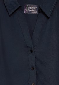 Camicia bottonata blu navy con colletto, caratterizzata da una texture liscia e una leggera lucentezza, include un'etichetta con il testo che indica il marchio e il design.