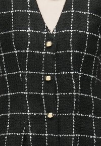 Čierna textúrovaná látka s bielou mriežkou a tromi lesklými zlatými gombíkmi na výstrihu do V.